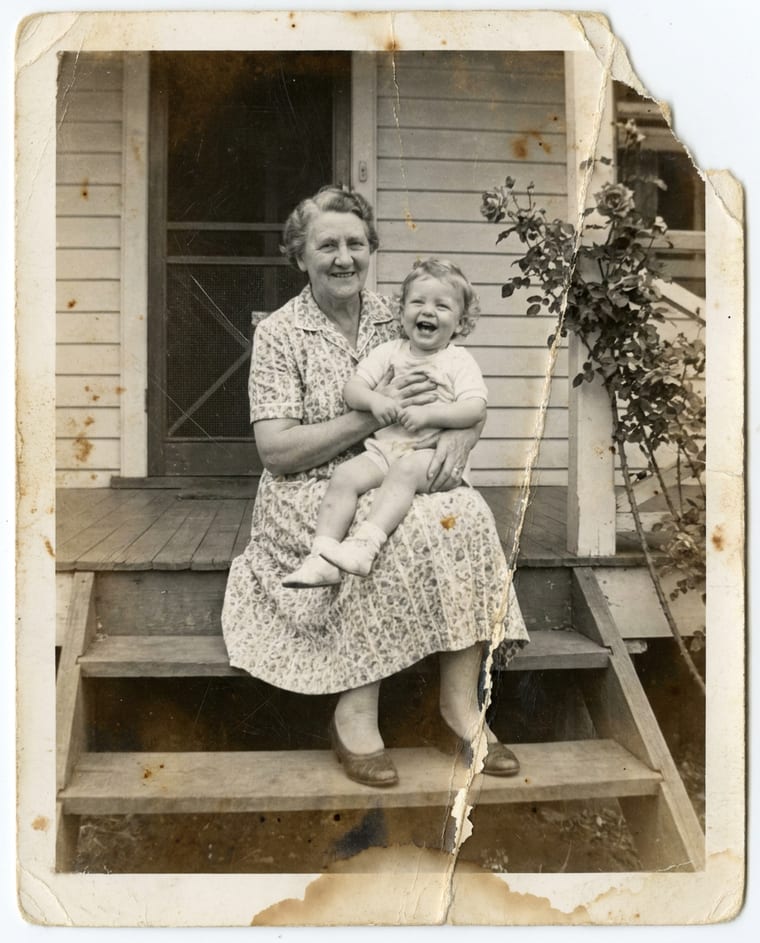 A 1958 black-and-white snapshot of a grandmother holding a laughing toddler on porch steps. The print has a torn corner, a long crease down the right side, brown foxing spots and a water stain.